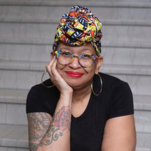 Claudia Love Mair, a Black woman wearing a colorful turban and glasses. She wears bright red lipstick and is leaning her head in her hand.