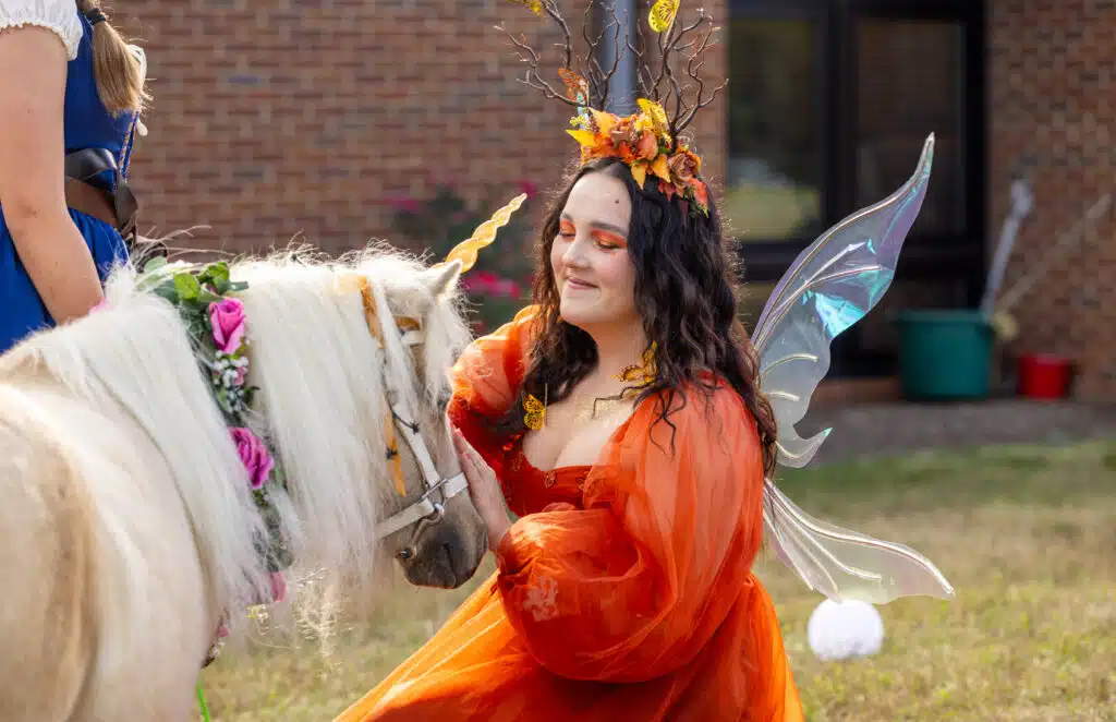 A woman cosplaying as a fall fairy pets a white pony with a unicorn horn.