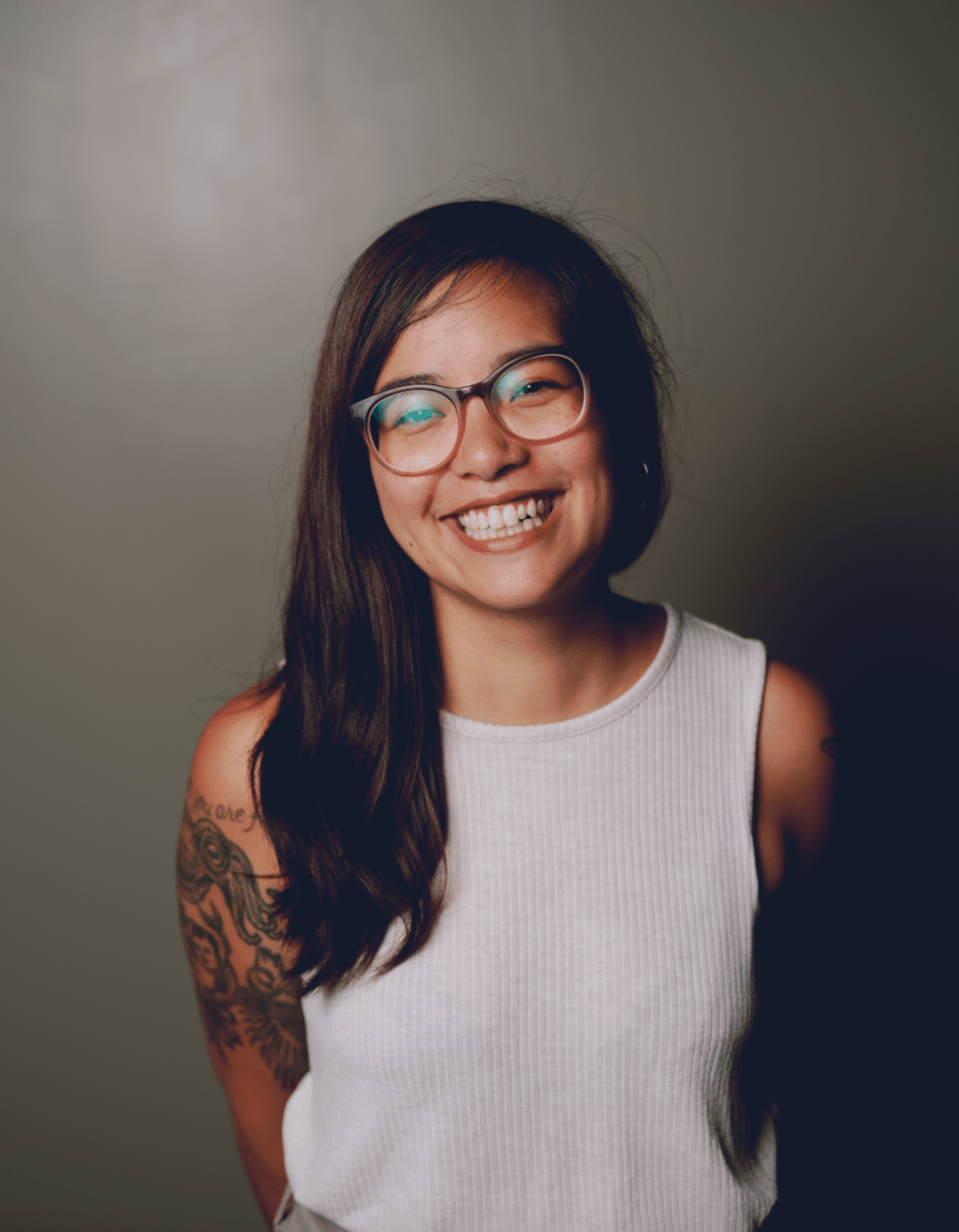 Photo of Danni Quintos, an Asian American woman with long brown hair. She wears glasses and is smiling. A large tattoo covers her right arm.