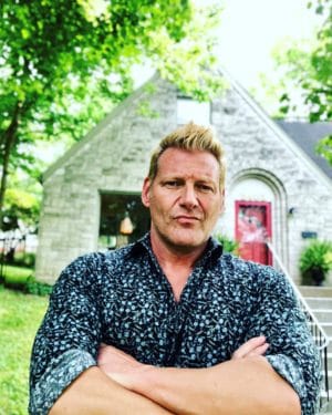 Author David Domine stands in front of a limestone house with a red door. He is a white man with blond hair, and he's wearing a shirt with a black print. His arms are crossed.