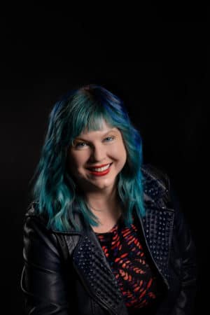 Author Gwenda Bond, a white woman with shoulder-length teal hair. She is smiling and wearing bright red lipstick.