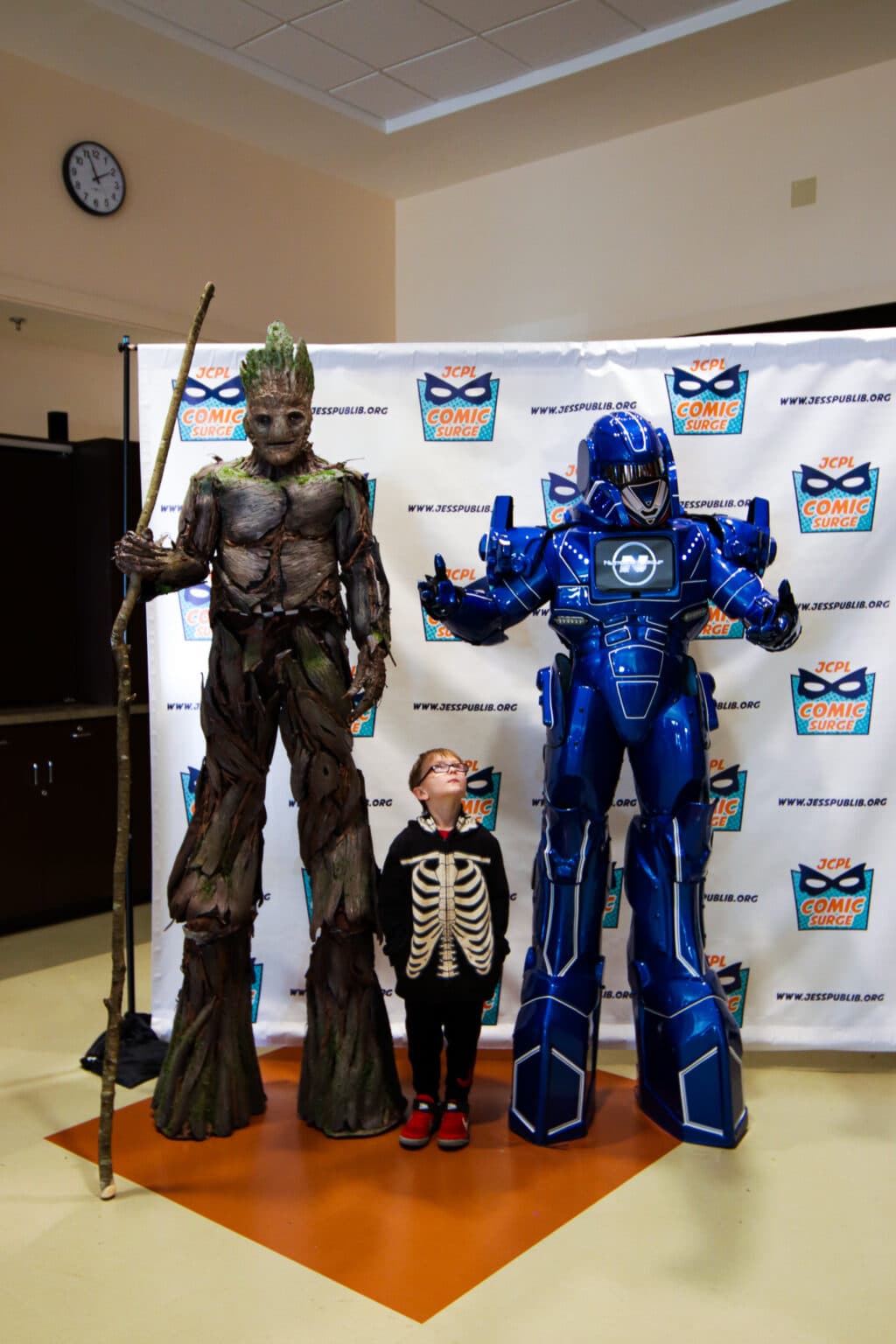 A small boy in a skeleton costume stands between Groot and the robot Big Blue. The boy looks up at Big Blue.