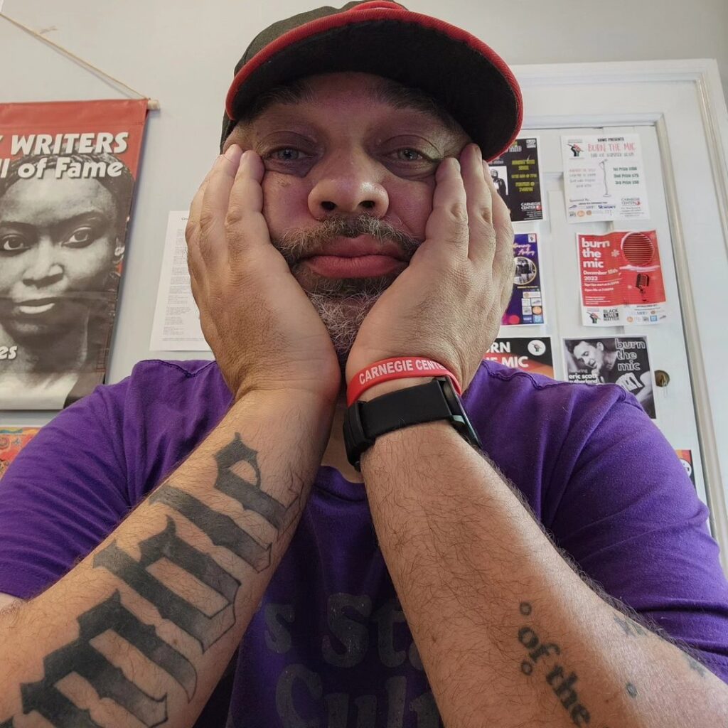 JC McPherson sits with his chin in his hands. He's wearing a baseball cap and a purple shirt. Behind him is a bulletin board with flyers for writing events.