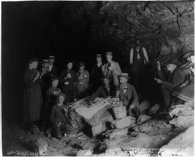 A group of people eat a picnic inside Mammoth Cave, circa 1892. Their meal is spread out on a cloth on top of a rock.