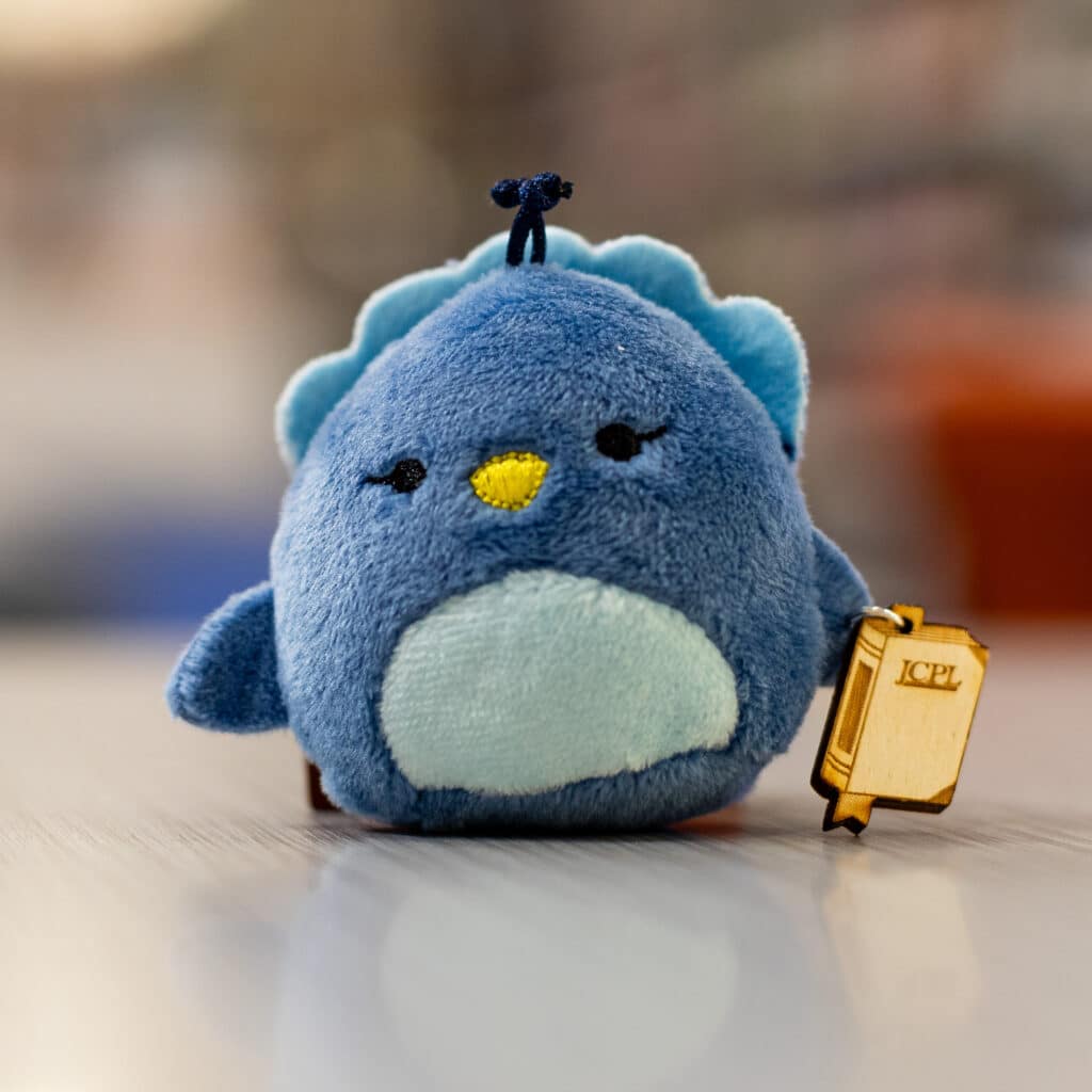 A mini blue Squishmallow holds a book made using JCPL's laser cutter.